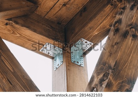 Close-up of two galvanized metal angle brackets securing vertical posts to horizontal beams in a stained timber frame structure.