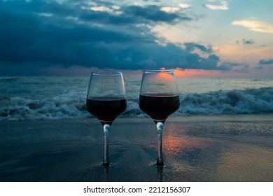 Close-up of two glasses of red wine standing in the sand against the background of the waves of the sea with a beautiful sunset. Beautiful picture of a romantic summer evening at the sea