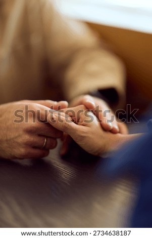 A close-up of two hands clasped over a dark wooden table, with a ring visible on one finger. Warm, intimate moment conveying support, trust, and connection in a casual dining setting.