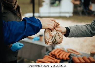 Close-up of two hands exchanging a brown paper bag at a market, representing a food purchase near a sausage stall. Trays of sausages are displayed in the background
