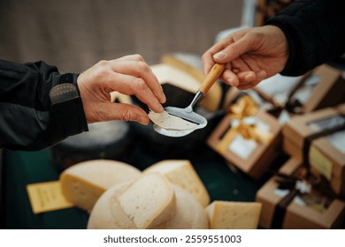 A close-up of two hands exchanging a cheese sample at a market. A vendor uses a wooden-handled slicer to serve a thin cheese slice to a customer, surrounded by cheese wheels and gift boxes - Powered by Shutterstock