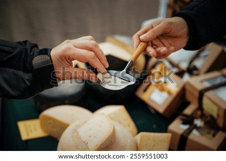 A close-up of two hands exchanging a cheese sample at a market. A vendor uses a wooden-handled slicer to serve a thin cheese slice to a customer, surrounded by cheese wheels and gift boxes