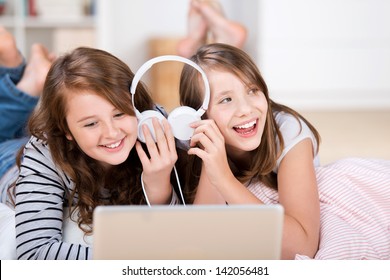 Close-up of two happy young teenage girls sharing headphones connected to a laptop to listen to music while laying on the living-room floor - Powered by Shutterstock