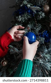 A close-up of two human hands decorating a frost-covered Christmas tree with a glittery blue bauble. The tree is covered in artificial snow and adorned with silver tinsel and blue accents.
