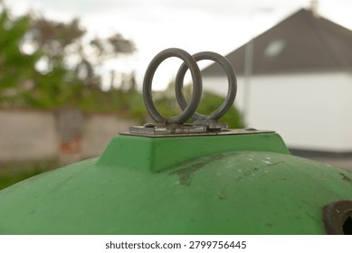 Close-up of two interlocked rusty metal lifting rings mounted on top of a rounded green industrial container or tank. The worn painted surface shows scratches