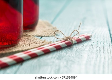 Closeup of two jars with organic raspberry juice, placed on a burlap coasters next to two striped red and white drinking straws tied together with a twine bow, placed on wooden boards surface