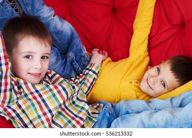 Close-up Of Two Peers On A Red Soft Coach. Two Kids Relaxing. Wearing Casual Clothes. Looking At The Camera And Smiling