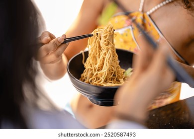 Close-up of two people enjoying a bowl of noodles. One person holds chopsticks, lifting noodles from the bowl, while the other also holds chopsticks, ready to eat. A relaxed, casual dining scene - Powered by Shutterstock