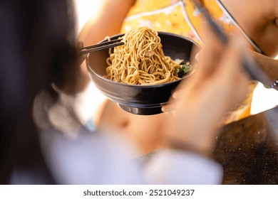 Close-up of two people enjoying a bowl of noodles. One person holds chopsticks, lifting noodles from the bowl, while the other also holds chopsticks, ready to eat. A relaxed, casual dining scene - Powered by Shutterstock