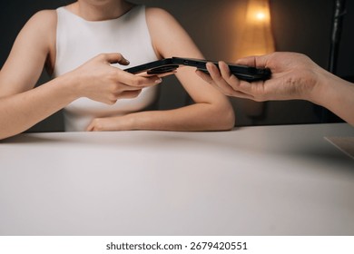 Close-up two people exchanging data using mobile phones sitting at white table at home. Man and woman comparing smartphone data, sharing digital information in casual workspace environment.