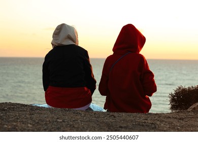 Close-up of two people in hooded jackets sitting on a cliff, watching the sunset over the ocean. Tranquil moment, ocean view, sunset, serene landscape, outdoor relaxation, coastal scenery - Powered by Shutterstock