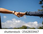 A close-up of two sporty individuals in sportswear and smartwatches giving each other a fist bump to celebrate reaching their running goal, with a nature and sky view in the background.