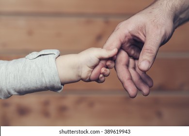 Closeup of two touching hands of small baby boy holding finger of male father as symbol of family love and trust on blurred wooden background, horizontal picture