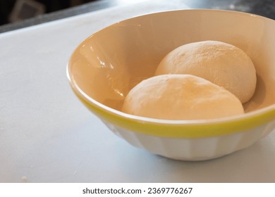 A close-up of two white round balls of bread dough in a white glass bowl with yellow trim. The bowl is on a white kitchen counter in a bakery.  The uncooked bread dough balls have a white flour coat. - Powered by Shutterstock