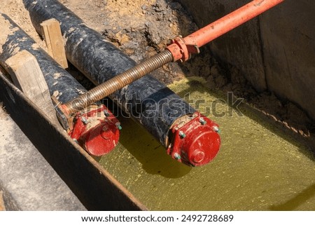 Close-up of an underground construction site with large black pipes and red connectors, secured with wooden supports and surrounded by muddy conditions.