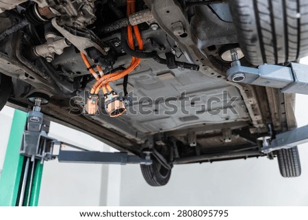 Close-up underside view of an electric vehicle lifted on a hydraulic service lift, revealing high-voltage orange cables and electrical connectors during maintenance inspection. Concept of EV engineering, battery system service, electric drivetrain, automotive diagnostics, vehicle repair, clean energy technology, and sustainable transportation.