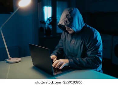 Close-up of an unidentifiable hacker with a hooded sweatshirt, working on a laptop in the kitchen. A portrayal of a dangerous and serious hacking scene. - Powered by Shutterstock