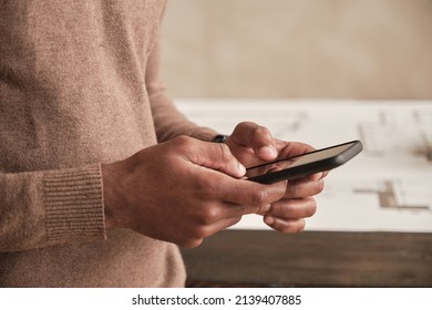 Close-up of unrecognizable Black architect in sweater standing against table with sketch and using internet on smartphone - Shutterstock ID 2139407885