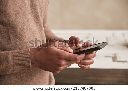 Close-up of unrecognizable Black architect in sweater standing against table with sketch and using internet on smartphone