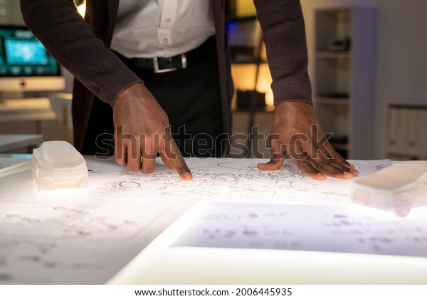 Close-up of\
unrecognizable black car engineer standing at table with 3D models\
in office and examining engine\
sketch
