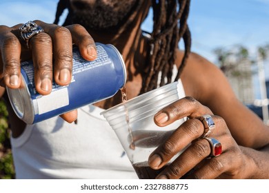 closeup of unrecognizable black ethnicity person with dreadlocks and rings on fingers outdoors at sunset pouring beer into plastic cup, copy space. - Powered by Shutterstock