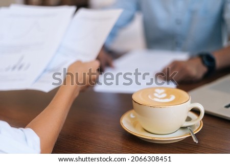 Close-up of unrecognizable businessman and businesswoman discussing and checking paper documents, sitting at table in cafe with cup of hot latte coffee with beautiful pattern, selective focus.