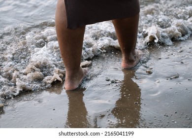 Closeup Unrecognizable Dark Skin Barefoot Woman Legs Walking Through Seaside, Beach, Foam Wash. Interracial Female On Vacation. Mixed Race People, Ethnicity, Diversity. Multicultural Person In Summer