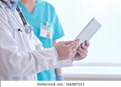 Close-up of unrecognizable doctor in lab coat using tablet while discussing online patients information with nurse in hospital