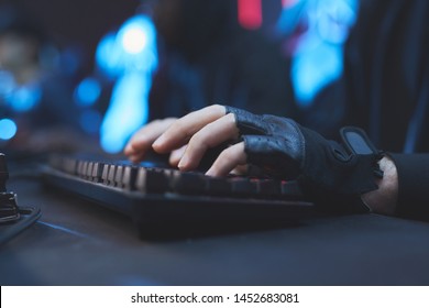 Close-up of unrecognizable hacker in black fingerless gloves sitting at desk and typing on computer keyboard while working on virus