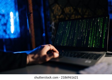 Close-up of unrecognizable hacker engaged in hacking into security system on laptop green binary code on screen with sitting at desk in dark basement room, with blue neon lights. - Powered by Shutterstock