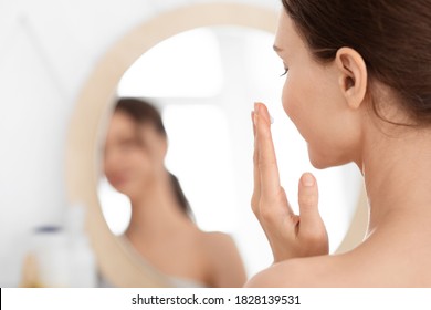 Closeup of unrecognizable lady using face cream at home, looking at mirror and smiling. Young woman applying beauty product on her cheeks, nourishing her sensitive skin after shower, face care