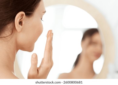 Closeup of unrecognizable lady using face cream at home, looking at mirror and smiling. Young woman applying beauty product on her cheeks, nourishing her sensitive skin after shower, face care