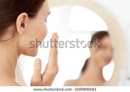 Closeup of unrecognizable lady using face cream at home, looking at mirror and smiling. Young woman applying beauty product on her cheeks, nourishing her sensitive skin after shower, face care