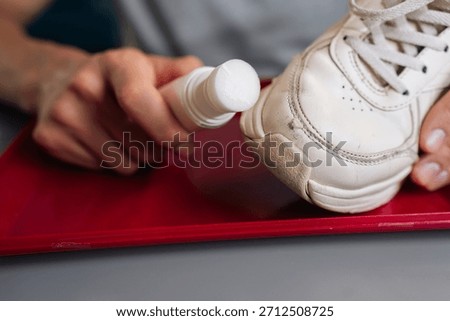 Close-up of unrecognizable man holding using bottle of shoe whitener and painting scuffed surface of dirty white sneaker, illustrating process of cleaning, recoloring, and restoring footwear.