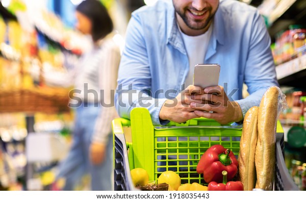 Closeup Of Unrecognizable Man Using Mobile Coupon\
App For Groceries Shopping Buying Food In Supermarket, Standing\
With Cart In Hypermarket. Smiling Guy Using Smartphone Purchasing\
Grocery In Shop