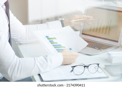 Close-up Of Unrecognizable Market Analyst Sitting At Desk And Comparing Graphs In Laptop And Papers While Working In Office, Behind Glass View