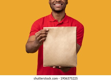 Closeup Of Unrecognizable Smiling African American Delivery Man In Red Uniform Holding And Giving Craft Paper Bag With Products From Restaurant To Camera, Isolated On Orange Studio Background