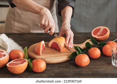 Close-up of unrecognizable woman in apron standing at dining table and cutting grapefruit with knife on wooden board