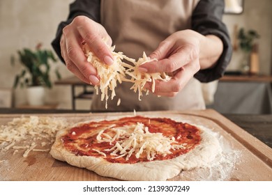 Close-up of unrecognizable woman standing at wooden board and sprinkling cheese on pizza dough with tomato sauce - Powered by Shutterstock