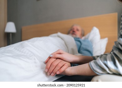Close-up of unrecognizable young woman child holding hand of sick aged father lying in hospital bed. Senior aged grandfather male patient resting at home and holding hand of granddaughter visitor.