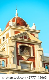 Close-up of the upper facade of a historic brick and stone building in Malaga, Spain, featuring a red tiled dome and multiple religious statues under a clear blue sky. Architectural details such as balconies, cornices, and a pediment are visible in warm sunlight.