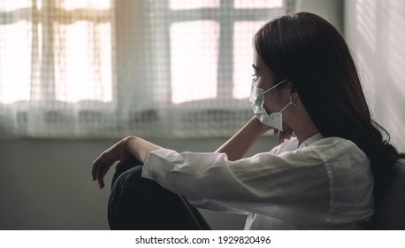 Close-up Of An Upset Asian Patient Woman Wearing A Face Mask At Home Or Hospital Alone On A Sofa Waiting For A Doctor. Hospital And Health Care During Coronavirus Or Covid-19 Quarantine Concept.