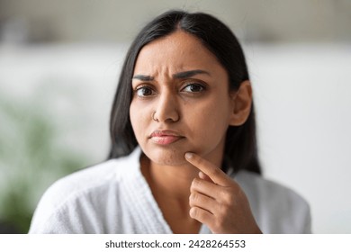 Closeup of upset young indian woman in white bathrobe looking at camera and touching her face at bathroom, suffering from skin problems. Acne, wrinkles, dull skin concept