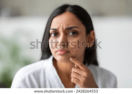 Closeup of upset young indian woman in white bathrobe looking at camera and touching her face at bathroom, suffering from skin problems. Acne, wrinkles, dull skin concept