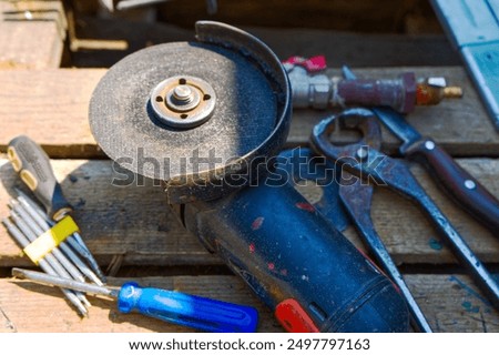 A close-up of a used angle grinder with a worn disc, alongside various hand tools like pliers and screwdrivers, all resting on a wooden surface, suggesting a workshop setting.