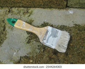 Close-up of a used, paint-stained brush with a wooden handle and green tip lying on a mossy, rough concrete surface, symbolizing painting, creativity, and DIY projects - Powered by Shutterstock