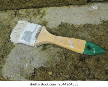 Close-up of a used, paint-stained brush with a wooden handle and green tip lying on a mossy, rough concrete surface, symbolizing painting, creativity, and DIY projects - Powered by Shutterstock