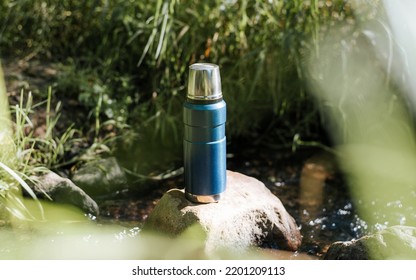 Close-up Of A Vacuum Thermos, A Camping Flask On A Stone Near A Stream In The Forest On A Sunny Day.