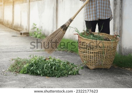 Closeup vellager holds stick broom to sweep pile of grass beside the street and put in the basket. Concept, community service, get rid of grass and garbage beside street.                           