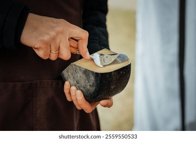 Close-up of a vendor slicing a wedge of artisan cheese with a wooden-handled slicer. The cheese features a textured black rind and creamy yellow interior, perfect for sampling - Powered by Shutterstock